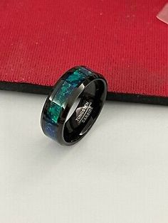 a black ring with blue and green marble inlays on it, sitting next to a red cloth