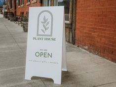 an open sign on the sidewalk in front of a brick building with a plant house logo