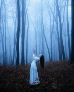 a woman in a white dress standing in the middle of a forest with her hands up