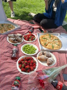 a woman sitting on the grass eating pizza and other food at an outdoor picnic table