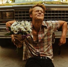 a man sitting on the ground with flowers in front of his face and holding a bouquet