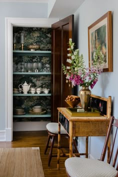 a dining room table and chairs with flowers in vases on the shelf next to it