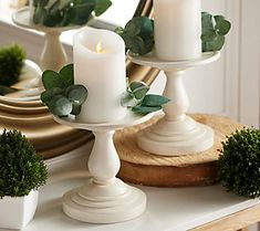 three white candles sitting on top of a table next to potted plants and a mirror