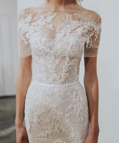 a woman wearing a white dress with sheer shoulders and flowers on the shoulder, standing in front of a wall