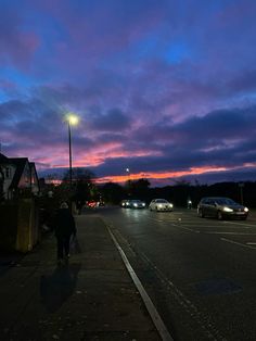 a person walking down the street at dusk with cars parked on the side walk and buildings in the background