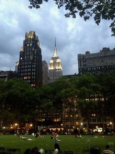 the city skyline is lit up at night with people sitting in the park and walking around