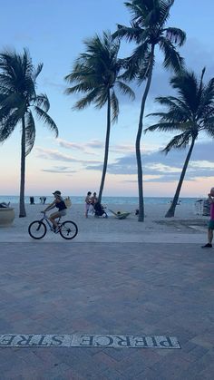 a man riding a bike down a sidewalk next to palm trees and people sitting on the beach