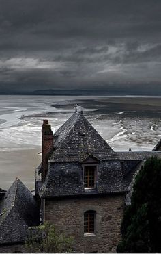 an old building on the beach with dark clouds in the sky above it and water below