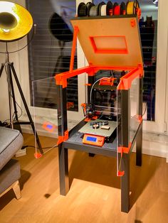 an orange and black 3d printer sitting on top of a wooden table next to a gray chair