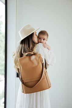 a woman holding a baby in her arms while wearing a tan purse and white dress