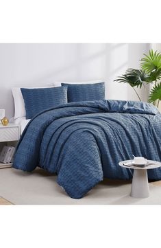 a bed with blue comforter and pillows in a room next to a plant on the floor