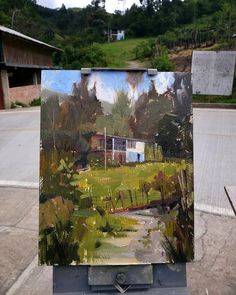 an easel with a painting on top of it in front of a building and trees