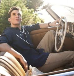 a man sitting in the driver's seat of an old car with his hand on the steering wheel