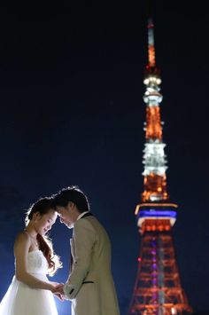 a bride and groom standing in front of the tokyo tower at night with their hands together