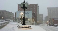 a clock on a pole in the middle of a snow covered street with buildings behind it