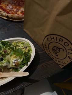 a salad in a paper bowl on a table next to a bag of chips and pizza