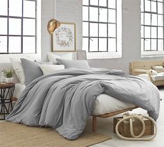 a bed with grey sheets and pillows in a room next to two large windows on the wall