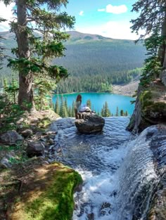 a person sitting on top of a rock next to a waterfall in the woods near a lake