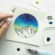 a person holding a pen and drawing with watercolors on paper next to some other items