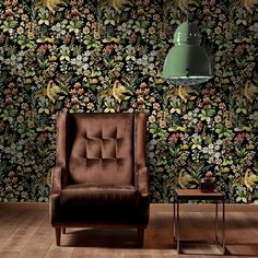 a brown chair sitting in front of a wall with flowers on it and a green lamp hanging from the ceiling