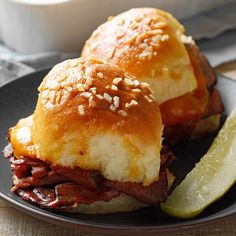 two pastrami sandwiches on a black plate next to a pickle and sauce