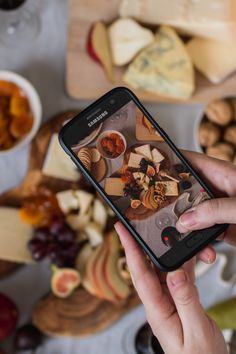 a person holding a cell phone in front of food on a table with cheeses and nuts