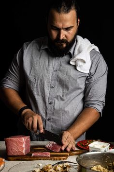 a man with a beard is cutting meat on a board in front of other foods