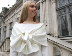 a woman standing in front of a building wearing a white top with a bow on it