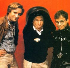 three men standing next to each other in front of a red wall wearing black leather jackets