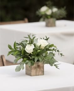 white roses in a wooden vase on a table with greenery and chairs behind them