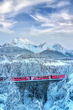 a red train traveling over a bridge in the middle of snow covered mountains and trees