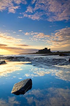 the sun is setting over an ocean with rocks in the foreground and a castle in the background