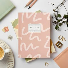 a pink notebook sitting on top of a white table next to a cup of coffee