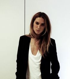 a woman standing in front of a white wall wearing a black blazer and jeans