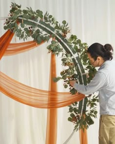 a woman is placing flowers on the top of an orange and white wedding arch with ribbon