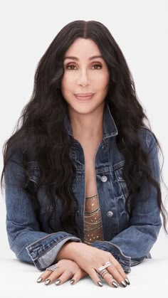 a woman with long black hair wearing a jean jacket and silver nails sitting on the floor