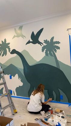 a woman sitting on the floor painting a dinosaur mural