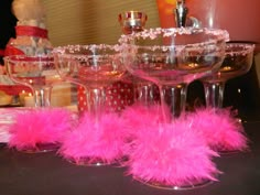 three wine glasses with pink feathers sit on a table next to other glassware and decorations