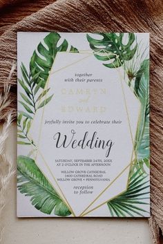 a wedding card with greenery and gold foil on it, sitting next to some feathers