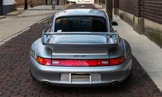 the back end of a silver sports car parked on a brick sidewalk next to a building