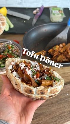 a person holding up a sandwich in front of some other food on a wooden table