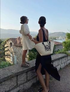 two women standing next to each other near a stone wall and looking at the water