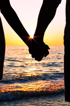 two people holding hands while standing in front of the ocean at sunset, with the sun setting behind them