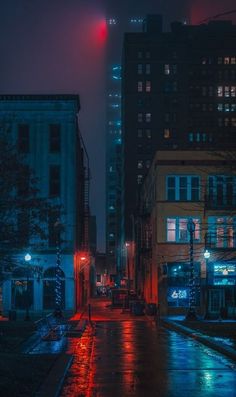 an empty city street at night with buildings lit up in the background and rain on the ground