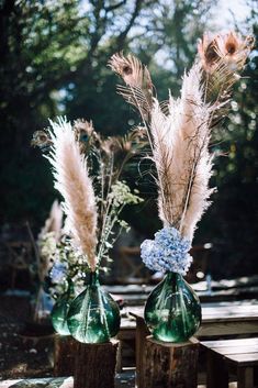 two vases filled with flowers and feathers on top of a wooden table in the woods