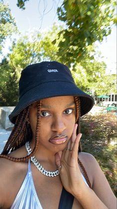 a woman wearing a black hat with braids on her head and holding her hand to her face