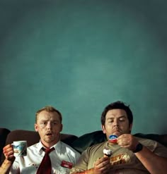 two men sitting on a couch eating cereal and drinking sodas while one man looks at the camera