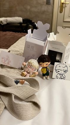 two figurines sitting on top of a bed next to an open gift box