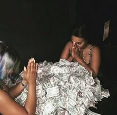 two women sitting at a table covered in money with one woman covering her face and the other