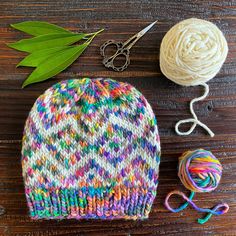 a knitted hat next to yarn and scissors on a wooden table with green leaves